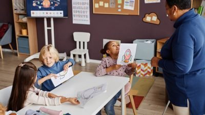Cute little learner of primary school boasting with her drawing of rocket while showing paper to African American teacher at lesson